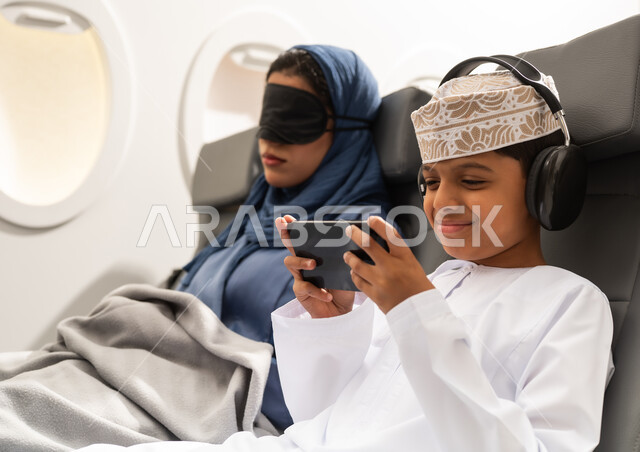 Oman Air, an Arab Gulf woman from Oman wearing an abaya and hijab sits in an airplane seat on a flight, wearing a black eye mask, while an Arab Gulf boy wearing headphones plays on his mobile phone with gestures of happiness and joy.