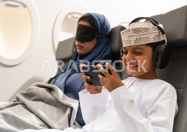 Gestures of happiness and joy, a family tourist trip through Oman Air, an Arab Gulf woman from Oman wearing an abaya and hijab sitting on an airplane seat during a flight, wearing a black eye mask and a travel pillow around her neck, an Arab Gulf boy wearing headphones and playing on a mobile phone.