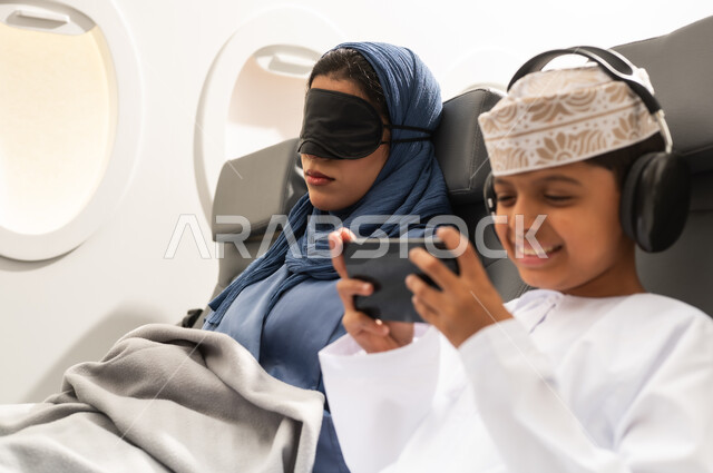 رحلة سياحية عائلية من خلال خطوط الطيران  العماني, امراة عربيه خليجيه عمانية ترتدي العباءة و الحجاب تجلس على كرسي الطائرة فى رحله جويه تضع الماسك الاسود على عينها ومخده السفرحول رقبتها, ولد عربي خليجي يرتدي السماعات و يقوم بالعب على الهاتف المحمول بايماءات السعادة و السرور