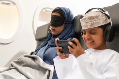رحلة سياحية عائلية من خلال خطوط الطيران  العماني, امراة عربيه خليجيه عمانية ترتدي العباءة و الحجاب تجلس على كرسي الطائرة فى رحله جويه تضع الماسك الاسود على عينها ومخده السفرحول رقبتها, ولد عربي خليجي يرتدي السماعات و يقوم بالعب على الهاتف المحمول بايماءات السعادة و السرور