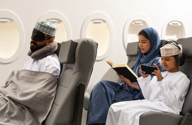 Spending the summer vacation with family, an Arab Gulf Omani woman wearing an abaya and hijab sits in an airplane seat on a flight reading a book alongside her son, an Arab Gulf boy wearing headphones and playing on a mobile phone with gestures of happiness and joy.