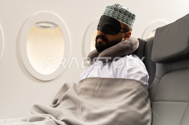 النوم والراحه والاسترخاء داخل الطائره, رجل عربي خليجي عماني يرتدي الدشداشه والكمه يجلس على كرسيه ويضع الماسك الاسود على عينه ومخده السفرحول رقبته, الطيران الخليجي العماني, مفهوم السياحة و السفر