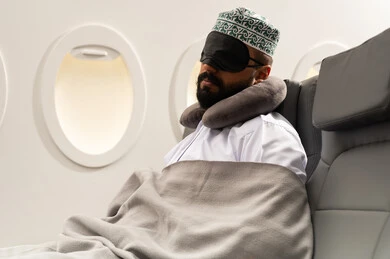 النوم والراحه والاسترخاء داخل الطائره, رجل عربي خليجي عماني يرتدي الدشداشه والكمه يجلس على كرسيه ويضع الماسك الاسود على عينه ومخده السفرحول رقبته, الطيران الخليجي العماني, مفهوم السياحة و السفر