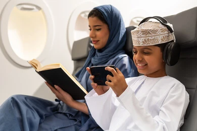 The concept of tourism and travel, spending enjoyable times during vacation, an Arab Gulf Omani woman wearing an abaya and hijab sitting in an airplane seat on a flight reading a book accompanied by her son, an Arab Gulf boy wearing headphones and playing on a mobile phone with gestures of happiness and joy.