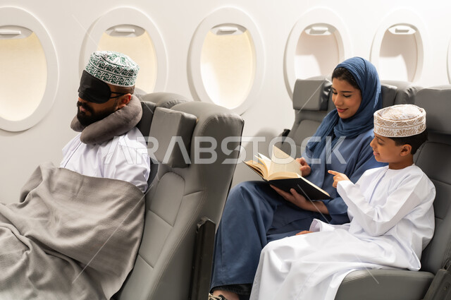 قضاء وقت ممتع اثناء السفر , امراة عربيه خليجيه عمانية ترتدي العباءة و الحجاب تجلس على كرسي الطائرة فى رحله جويه تقوم بقراءة كتاب برفقة ابنها , رجل خليجي يرتدي  الدشداشة و الكمه يضع ماسك اسود على عينيه و وسادة النوم , رحله سياحية عائلية