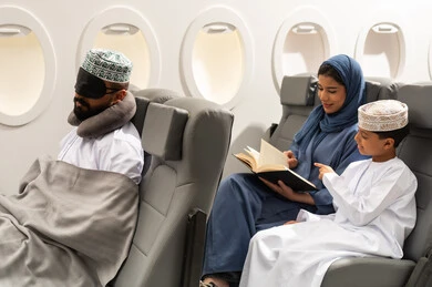 Spending enjoyable time while traveling, an Arab Gulf woman from Oman wearing an abaya and hijab sits on an airplane seat during a flight reading a book accompanied by her son, a Gulf man wearing a dishdasha and a ghutrah puts a black mask over his eyes and a sleep pillow, a family tourist trip.