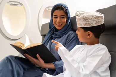 رحلة سياحية عائلية، ولد يقضي وقت الرحلة برفقة والدته يقوم بالاشارة الى شي ما، امرأة عربية خليجية عمانية ترتدي العباءة و الحجاب تمسك بيديها كتاب، قضاء العطلة الصيفية برفقة العائلة، خطوط الطيران العماني, ايماءات السعادة و السرور
