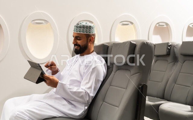 متابعة الأفلام والمسلسلات الممتعة, استخدام التكنولوجيا والتقنيات الحديثة, تصفح مواقع التواصل الاجتماعي، المحادثة مع الأهل والأصدقاء، رجل عربي خليجي عماني يرتدي الدشداشة و الكمة يجلس على كرسي الطائرة فى رحله جوية يحمل بيده جهاز التابلت, ايماءات السعادة و السرور