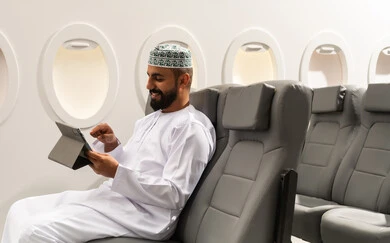 متابعة الأفلام والمسلسلات الممتعة, استخدام التكنولوجيا والتقنيات الحديثة, تصفح مواقع التواصل الاجتماعي، المحادثة مع الأهل والأصدقاء، رجل عربي خليجي عماني يرتدي الدشداشة و الكمة يجلس على كرسي الطائرة فى رحله جوية يحمل بيده جهاز التابلت, ايماءات السعادة و السرور