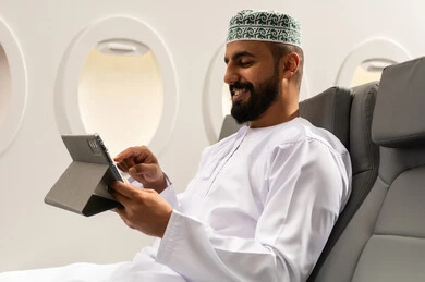 Browsing social media, using modern technology and techniques, following enjoyable movies and series, chatting with family and friends, an Arab Gulf Omani man wearing a dishdasha and kumma sitting on an airplane seat during a flight holding a tablet in his hand, gestures of happiness and joy.