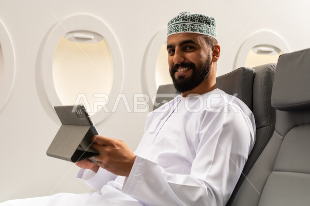 استخدام التكنولوجيا والتقنيات الحديثة, تصفح مواقع التواصل الاجتماعي، متابعة الأفلام والمسلسلات الممتعة، المحادثة مع الأهل والأصدقاء، رجل عربي خليجي عماني يرتدي الدشداشة و الكمة يجلس على كرسي الطائرة فى رحله جوية يحمل بيده جهاز التابلت, ايماءات السعادة و السرور