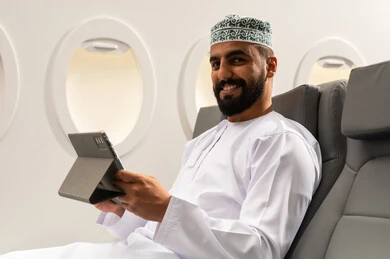 استخدام التكنولوجيا والتقنيات الحديثة, تصفح مواقع التواصل الاجتماعي، متابعة الأفلام والمسلسلات الممتعة، المحادثة مع الأهل والأصدقاء، رجل عربي خليجي عماني يرتدي الدشداشة و الكمة يجلس على كرسي الطائرة فى رحله جوية يحمل بيده جهاز التابلت, ايماءات السعادة و السرور