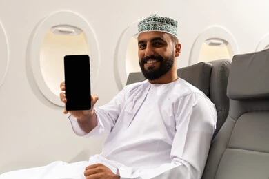 الترفيه والتسليه في اوقات الفراغ, تصفح وسائل التواصل الاجتماعي, استخدام جهاز تقني حديث ومتطور, رجل عربي خليجي عماني يرتدي الدشداشة والكمة يمسك بيده الهاتف المحمول جالسا على كرسيه داخل الطائره الخليجيه يقوم بعرض شاشة سوداء, الطيران الخليجي العماني, ايماءات السعادة و السرور
