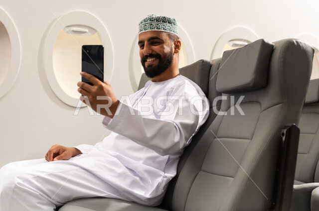 دمج التقنيات الحديثة فى الحياة اليومية, الاطمئنان على الاهل والاصدقاء ,اجراء مكالمة فيديو، تصفح مواقع التواصل الاجتماعي , رجل عربي خليجي عماني يرتدي الدشداشة و الكمة يجلس على كرسي الطائره في رحله جويه يقوم باستخدام الهاتف المحمول, ايماءات السعادة و السرور