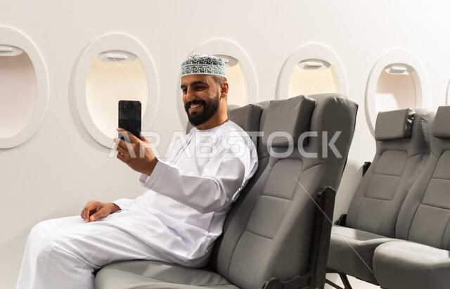 اجراء مكالمة فيديو, الاطمئنان على الاهل والاصدقاء ، تصفح مواقع التواصل الاجتماعي , رجل عربي خليجي عماني يرتدي الدشداشة و الكمة يجلس على كرسي الطائره في رحله جويه يقوم باستخدام الهاتف المحمول, دمج التقنيات الحديثة فى الحياة اليومية, ايماءات السعادة و السرور