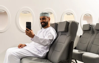 اجراء مكالمة فيديو, الاطمئنان على الاهل والاصدقاء ، تصفح مواقع التواصل الاجتماعي , رجل عربي خليجي عماني يرتدي الدشداشة و الكمة يجلس على كرسي الطائره في رحله جويه يقوم باستخدام الهاتف المحمول, دمج التقنيات الحديثة فى الحياة اليومية, ايماءات السعادة و السرور