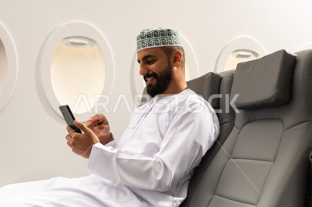 الاطمئنان على الاهل والاصدقاء ، تصفح مواقع التواصل الاجتماعي , رجل عربي خليجي عماني يرتدي الدشداشة و الكمة يجلس على كرسي الطائره في رحله جويه يقوم باستخدام الهاتف المحمول, دمج التقنيات الحديثة فى الحياة اليومية, ايماءات السعادة و السرور