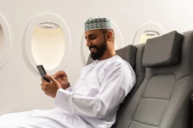 الاطمئنان على الاهل والاصدقاء ، تصفح مواقع التواصل الاجتماعي , رجل عربي خليجي عماني يرتدي الدشداشة و الكمة يجلس على كرسي الطائره في رحله جويه يقوم باستخدام الهاتف المحمول, دمج التقنيات الحديثة فى الحياة اليومية, ايماءات السعادة و السرور