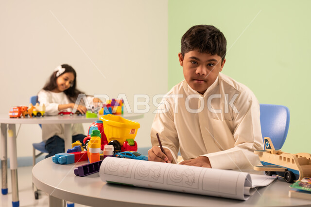 مفهوم الاهتمام بالهوايات،قضاء اوقات ممتعه ، ولد عماني مصاب بمتلازمه داون يقوم بالرسم والتلوين ، تنمية المهارات الفنيه في الروضات السعودية  ،بنت عربيه سعوديه ترتدي الزي الكاجوال تجلس على كرسي متحرك، تلعب بالالعاب البلاستيكيه الملونه