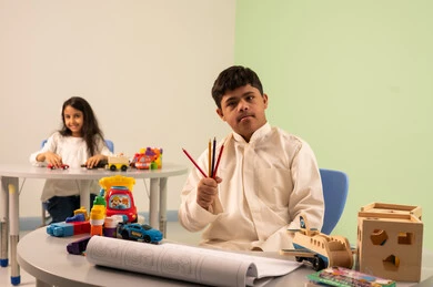 بنت عربيه سعوديه ترتدي الزي الكاجوال تجلس على كرسي متحرك، تلعب بالالعاب البلاستيكيه الملونه، مفهوم الاهتمام بالهوايات،تنمية المهارات الفنيه في الروضات السعودية  ،ولد عماني مصاب بمتلازمه داون ينظر الى الكاميرا بايماءات السعاده،قضاء اوقات ممتعه