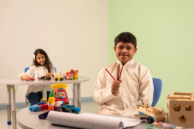 Developing artistic skills in Saudi kindergartens, an Omani boy with Down syndrome looks at the camera with happy gestures, a Saudi Arabian girl wearing casual clothes sitting in a wheelchair, playing with colorful plastic toys, having fun, the concept of interest in hobbies