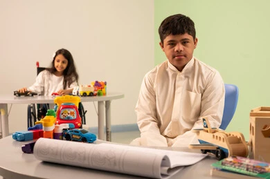 Having fun, the concept of interest in hobbies, developing artistic skills in Saudi kindergartens, an Omani boy with Down syndrome looks at the camera with happy gestures, a Saudi Arabian girl wearing casual clothes sitting on a wheelchair, playing with colorful plastic toys