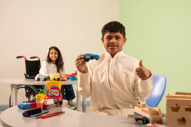 Saudi society's interest in people with disabilities, encouraging, caring for and developing children's skills, creativity and developing mental and intellectual skills in Saudi kindergartens, a Saudi Arabian girl wearing casual clothes sitting in a wheelchair, assembling blocks with happy expressions, recreational activities for people with special needs, an Omani boy with Down syndrome playing with colorful plastic toys