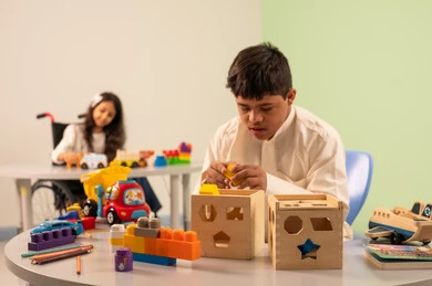 تشجيع ورعاية وتطوير مهارات الأطفال،نشاطات ترفيهية لذوي الاحتياجات الخاصة ،الابداع وتنمية المهارات الذهنيه والفكريه في الروضات السعودية ،  اهتمام المجتمع السعودي بأصحاب الهمم, بنت عربيه سعوديه ترتدي الزي الكاجوال تجلس على كرسي متحرك، تقوم باللعب بالالعاب البلاستيكيه الملونه  بتعابير السعاده ،ولد عماني مصاب بمتلازمه داون يقوم باللعب بالالعاب الخشبيه