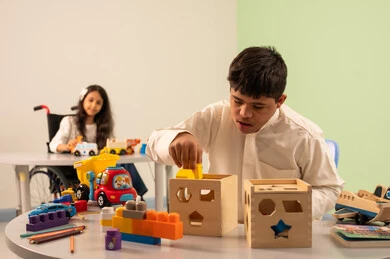 بنت عربيه سعوديه ترتدي الزي الكاجوال تجلس على كرسي متحرك، تقوم باللعب بالالعاب البلاستيكيه الملونه  بتعابير السعاده ، تشجيع ورعاية وتطوير مهارات الأطفال،نشاطات ترفيهية لذوي الاحتياجات الخاصة ،الابداع وتنمية المهارات الذهنيه والفكريه في الروضات السعودية ،  اهتمام المجتمع السعودي بأصحاب الهمم, ولد عماني مصاب بمتلازمه داون يقوم باللعب بالالعاب الخشبيه