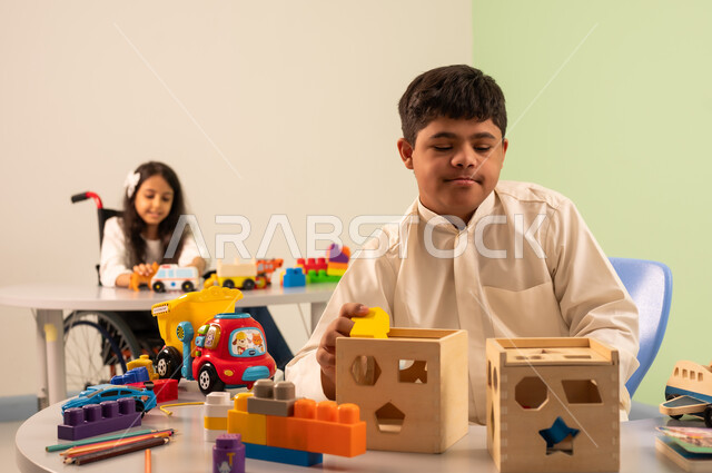 الابداع وتنمية المهارات الذهنيه والفكريه في الروضات السعودية ،   بنت عربيه سعوديه ترتدي الزي الكاجوال تجلس على كرسي متحرك، تقوم باللعب بالالعاب البلاستيكيه الملونه  بتعابير السعاده ، تشجيع ورعاية وتطوير مهارات الأطفال،نشاطات ترفيهية لذوي الاحتياجات الخاصة ،اهتمام المجتمع السعودي بأصحاب الهمم, ولد عماني مصاب بمتلازمه داون يقوم باللعب بالالعاب الخشبيه