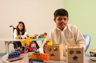 الابداع وتنمية المهارات الذهنيه والفكريه في الروضات السعودية ،   بنت عربيه سعوديه ترتدي الزي الكاجوال تجلس على كرسي متحرك، تقوم باللعب بالالعاب البلاستيكيه الملونه  بتعابير السعاده ، تشجيع ورعاية وتطوير مهارات الأطفال،نشاطات ترفيهية لذوي الاحتياجات الخاصة ،اهتمام المجتمع السعودي بأصحاب الهمم, ولد عماني مصاب بمتلازمه داون يقوم باللعب بالالعاب الخشبيه