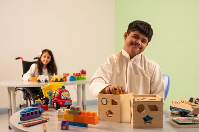 نشاطات ترفيهية لذوي الاحتياجات الخاصة ، الابداع وتنمية المهارات الذهنيه والفكريه في الروضات السعودية ، بنت عربيه سعوديه ترتدي الزي الكاجوال تجلس على كرسي متحرك، تقوم باللعب بالالعاب البلاستيكيه الملونه  بتعابير السعاده ، تشجيع ورعاية وتطوير مهارات الأطفال،اهتمام المجتمع السعودي بأصحاب الهمم, ولد عماني مصاب بمتلازمه داون يقوم باللعب بالالعاب الخشبيه