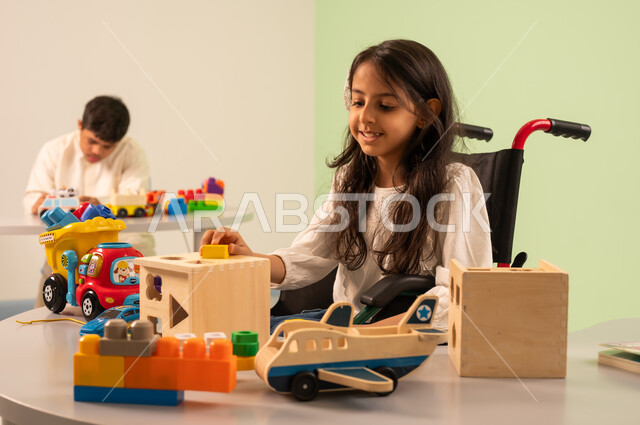 Saudi society's interest in people of determination, recreational activities for people with special needs, creativity and development of mental and intellectual skills in Saudi kindergartens, a Saudi Arabian girl wearing casual clothes sitting in a wheelchair, playing with wooden toys with happy expressions, encouraging, caring for and developing children's skills, an Omani boy with Down syndrome playing with colorful plastic toys