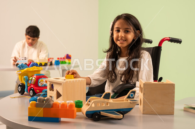 Encouraging, nurturing and developing children's skills, Saudi society's interest in people of determination, recreational activities for people with special needs, creativity and development of mental and intellectual skills in Saudi kindergartens, a Saudi Arabian girl wearing casual clothes sitting in a wheelchair, playing with wooden toys with happy expressions, an Omani boy with Down syndrome playing with colorful plastic toys