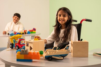 A Saudi Arabian girl wearing casual clothes sitting on a wheelchair, playing with wooden toys with happy expressions, encouraging, caring for and developing children's skills, Saudi society's interest in people with disabilities, recreational activities for people with special needs, creativity and development of mental and intellectual skills in Saudi kindergartens, an Omani boy with Down syndrome playing with colorful plastic toys