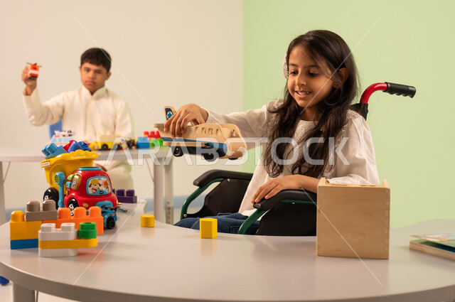 Saudi society's interest in people of determination, recreational activities for people with special needs, a Saudi Arabian girl wearing casual clothes sitting in a wheelchair, enjoying games with happy expressions, encouraging, caring for and developing children's skills, creativity and developing mental and intellectual skills in Saudi kindergartens, an Omani boy with Down syndrome playing with colorful plastic toys