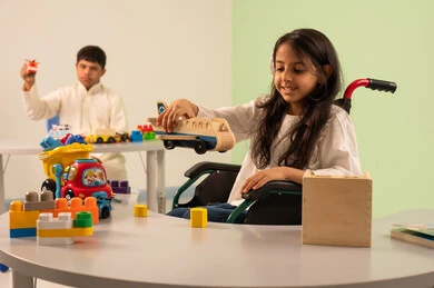 Saudi society's interest in people of determination, recreational activities for people with special needs, a Saudi Arabian girl wearing casual clothes sitting in a wheelchair, enjoying games with happy expressions, encouraging, caring for and developing children's skills, creativity and developing mental and intellectual skills in Saudi kindergartens, an Omani boy with Down syndrome playing with colorful plastic toys