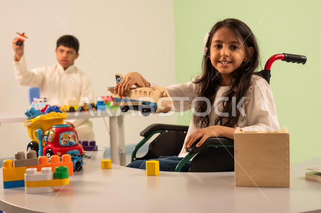 Creativity and development of mental and intellectual skills in Saudi kindergartens, Saudi society's interest in people of determination, recreational activities for people with special needs, a Saudi Arabian girl wearing casual clothes sitting in a wheelchair, enjoying games with happy expressions, encouraging, caring for and developing children's skills, an Omani boy with Down syndrome playing with colorful plastic toys