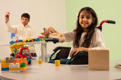 Creativity and development of mental and intellectual skills in Saudi kindergartens, Saudi society's interest in people of determination, recreational activities for people with special needs, a Saudi Arabian girl wearing casual clothes sitting in a wheelchair, enjoying games with happy expressions, encouraging, caring for and developing children's skills, an Omani boy with Down syndrome playing with colorful plastic toys