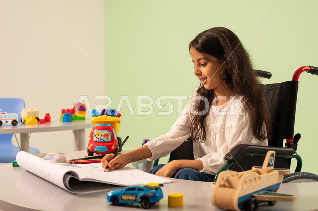 قضاء اوقات ممتعه ، تنمية المهارات الفنيه في الروضات السعودية ، مفهوم الاهتمام بالهوايات،بنت عربيه سعوديه ترتدي الزي الكاجوال تجلس على كرسي متحرك، تقوم بالرسم والتلوين بتعابير التركيز والاستمتاع