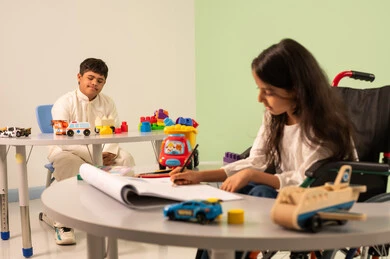 Omani boy with Down syndrome looking at the camera with happy gestures, developing artistic skills in Saudi kindergartens, having fun, the concept of interest in hobbies, Saudi Arabian girl wearing casual clothes sitting in a wheelchair, drawing and coloring with expressions of concentration and enjoyment