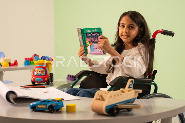 Promoting mental activity, the concept of interest in hobbies, developing mental and intellectual skills in Saudi kindergartens, spending enjoyable times, a Saudi Arabian girl wearing casual clothes sitting in a wheelchair, reading colorful educational books with happy expressions