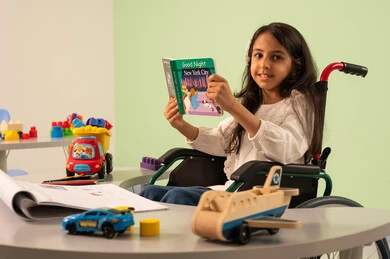 تعزيز النشاط الذهني ، مفهوم الاهتمام بالهوايات، تنمية المهارات الذهنيه والفكريه في الروضات السعودية ،  قضاء اوقات ممتعه ، بنت عربيه سعوديه ترتدي الزي الكاجوال تجلس على كرسي متحرك، تقوم بقراءه الكتب التعليميه الملونه بتعابير السعاده