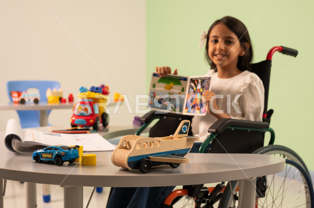 تعزيز النشاط الذهني ،  قضاء اوقات ممتعه ،  تنمية المهارات الذهنيه والفكريه في الروضات السعودية ، مفهوم هوايه المطالعه ، بنت عربيه سعوديه ترتدي الزي الكاجوال تجلس على كرسي متحرك، تقوم بعرض الكتاب التعليمي الملون بتعابير السعاده