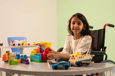Creativity and development of mental and intellectual skills in Saudi kindergartens, encouraging, nurturing and developing children's skills, recreational activities for people with special needs, Saudi society's interest in people of determination, spending enjoyable times, a Saudi Arabian girl wearing casual clothes sitting in a wheelchair, assembling blocks with happy expressions