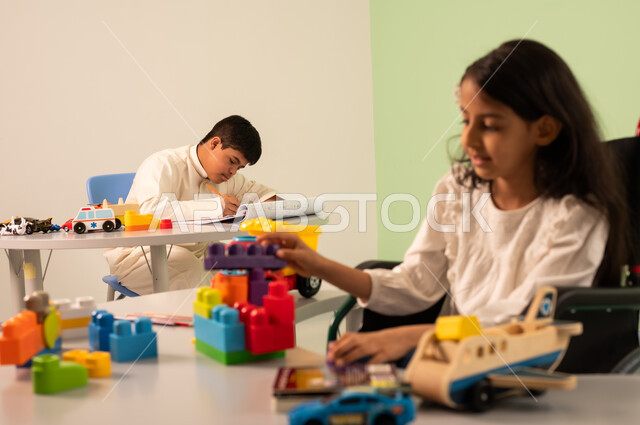 Encouraging, nurturing and developing children's skills, creativity and developing mental and intellectual skills in Saudi kindergartens, recreational activities for people with special needs, Saudi society's interest in people of determination, a Saudi Arabian girl wearing casual clothes sitting in a wheelchair, assembling blocks with happy expressions, an Omani boy with Down syndrome drawing and coloring
