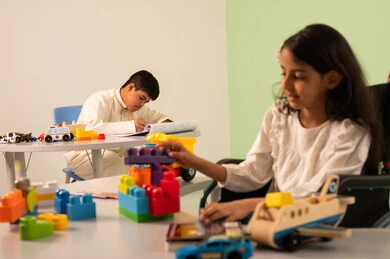 تشجيع ورعاية وتطوير مهارات الأطفال،  الابداع وتنمية المهارات الذهنيه والفكريه في الروضات السعودية ، نشاطات ترفيهية لذوي الاحتياجات الخاصة ، اهتمام المجتمع السعودي بأصحاب الهمم, بنت عربيه سعوديه ترتدي الزي الكاجوال تجلس على كرسي متحرك، تقوم بتركيب المكعبات بتعابير السعاده ،ولد عماني مصاب بمتلازمه داون يقوم بالرسم والتلوين