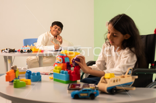 Recreational activities for people with special needs, Saudi society's interest in people of determination, encouraging, caring for and developing children's skills, creativity and developing mental and intellectual skills in Saudi kindergartens, a Saudi Arabian girl wearing casual clothes sitting in a wheelchair, assembling blocks with happy expressions, an Omani boy with Down syndrome drawing and coloring