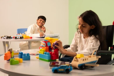 نشاطات ترفيهية لذوي الاحتياجات الخاصة ، اهتمام المجتمع السعودي بأصحاب الهمم, تشجيع ورعاية وتطوير مهارات الأطفال،  الابداع وتنمية المهارات الذهنيه والفكريه في الروضات السعودية ،بنت عربيه سعوديه ترتدي الزي الكاجوال تجلس على كرسي متحرك، تقوم بتركيب المكعبات بتعابير السعاده ،ولد عماني مصاب بمتلازمه داون يقوم بالرسم والتلوين