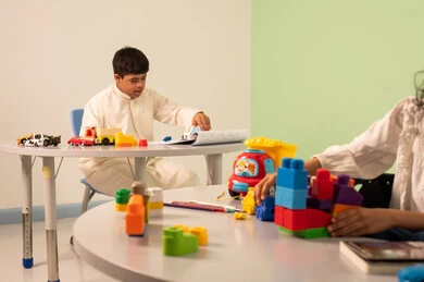 Close-up of a Saudi Arabian girl's hand wearing casual clothes sitting on a wheelchair, assembling blocks with happy expressions, recreational activities for people with special needs, Saudi society's interest in people of determination, encouraging, caring for and developing children's skills, creativity and developing mental and intellectual skills in Saudi kindergartens, an Omani boy with Down syndrome drawing and coloring